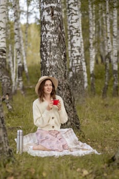 A young beautiful woman in a dress and a round hat reads a book outdoors in the forest and drinks tea. Romantic and vintage photo of a beautiful girl. Reading and relaxation