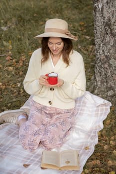 A young beautiful woman in a dress and a round hat reads a book outdoors in the forest and drinks tea. Romantic and vintage photo of a beautiful girl. Reading and relaxation