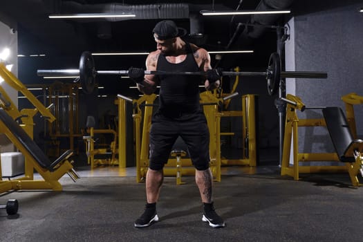 Attractive, tattooed, bearded sportsman in black sport gloves, shorts, vest, cap and sneakers. He is lifting a barbell, training his muscles, posing in dark gym with yellow equipment. Full length