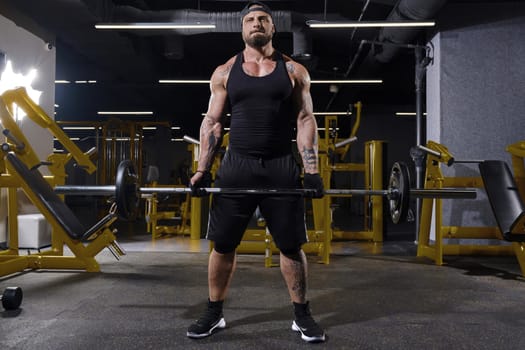 Young, tattooed, bearded athlete in black sport gloves, shorts, vest, cap and sneakers. He is lifting a barbell, training his muscles, posing in dark gym with yellow equipment. Full length