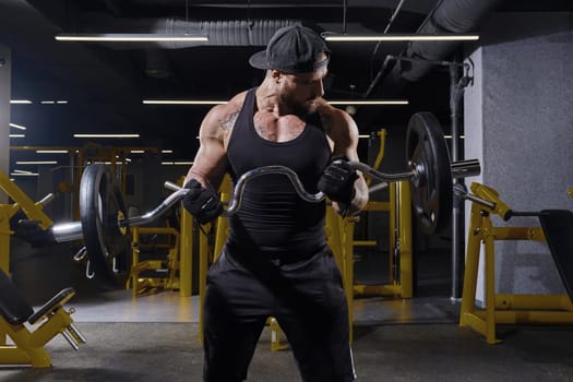 Tattooed, bearded sportsman in black sport gloves, shorts, vest and cap. He is lifting a barbell, training his biceps, looking at his muscles while posing in dark gym with yellow equipment. Close up