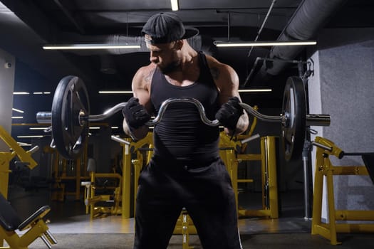 Tattooed, bearded male in black sport gloves, shorts, vest and cap. He is lifting a barbell, training his biceps, looking at his muscles while standing in dark gym with yellow equipment. Close up