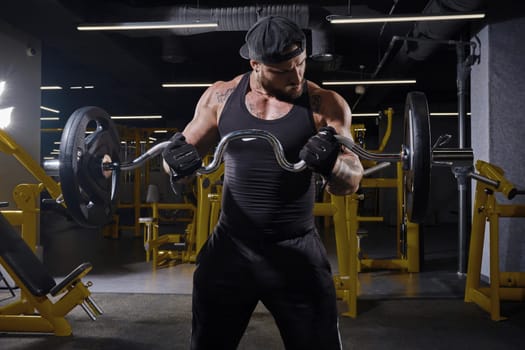 Tattooed, bearded man in black sport gloves, shorts, vest and cap. He is lifting a barbell, training his muscles, looking at his biceps while standing in dark gym with yellow equipment. Close up