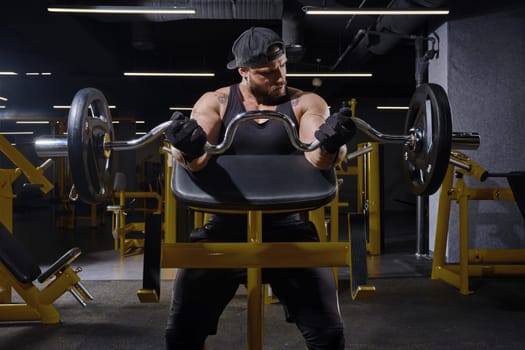 Tattooed, bearded athlete in black sport gloves, shorts, vest and cap. He is lifting a barbell, training his biceps while sitting on preacher curl bench at dark gym with yellow equipment. Close up