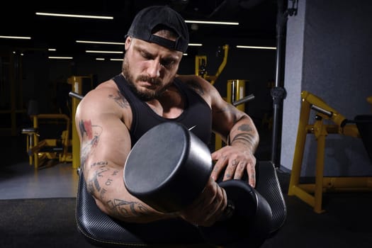 Tattooed, bearded guy in black vest and cap. He is lifting a dumbbell, training his biceps while sitting on preacher curl bench at dark gym with yellow equipment. Sport, fitness, lifestyle. Close up