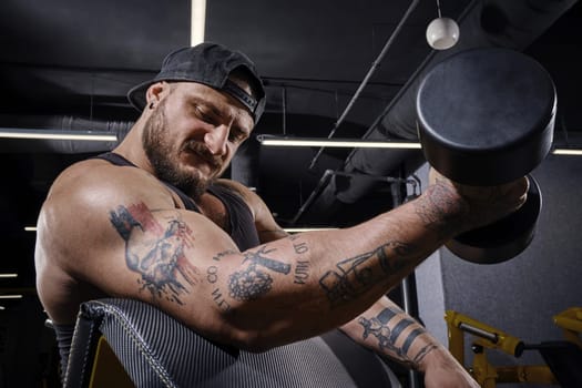 Tattooed, handsome bearded male in black vest and cap. He lifting dumbbell, training his biceps, sitting on preacher curl bench at dark gym with yellow equipment. Sport, fitness. Close up, side view