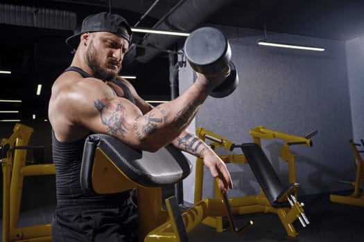 Tattooed, bearded athlete in black vest and cap. He is lifting a dumbbell, training his biceps, sitting on preacher curl bench at dark gym with yellow equipment. Sport, fitness. Close up, side view