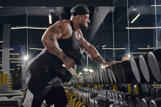 Young, tattooed, bearded muscular man in black shorts, vest, cap. Exercising with dumbbells for training his triceps, leaning on set of black weights in dark gym. Sport, fitness. Close up, side view