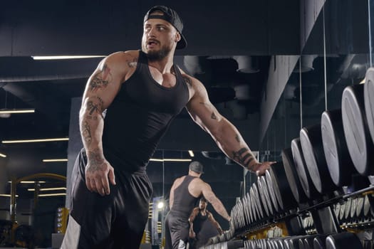 Young, tattooed, bearded male in black shorts, vest, cap. Going to do exercises with dumbbells for training muscles, leaning on set of black weights in dark gym. Sport, fitness. Close up, side view