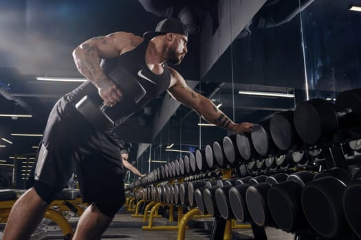 Tattooed, bearded muscular guy in black shorts, vest, cap. Exercising with dumbbells for training his triceps, leaning on set of black weights in dark gym. Sport, fitness. Close up, side view