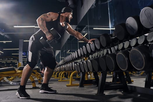 Tattooed, muscular sportsman in black shorts, vest, cap. Exercising with dumbbells for training his triceps, leaning on set of black weights in dark gym, looking at the mirror. Close up, side view