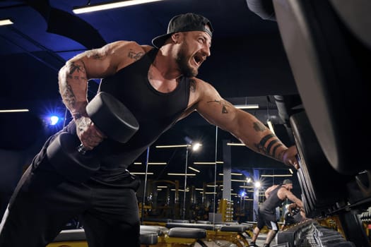 Handsome, tattooed, bearded muscular man in black shorts, vest, cap. Exercising with dumbbells for training his triceps, leaning on set of black weights. Dark gym. Sport, fitness. Close up, side view