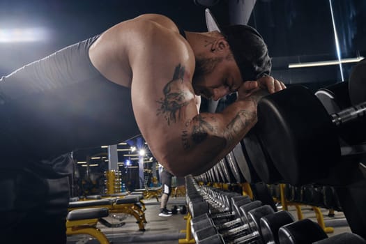Handsome, tattooed, bearded muscular athlete in black shorts, vest, cap. Having rest leaning on set of black dumbbells by his head and hands. Dark gym. Sport, fitness, lifestyle. Close up, side view