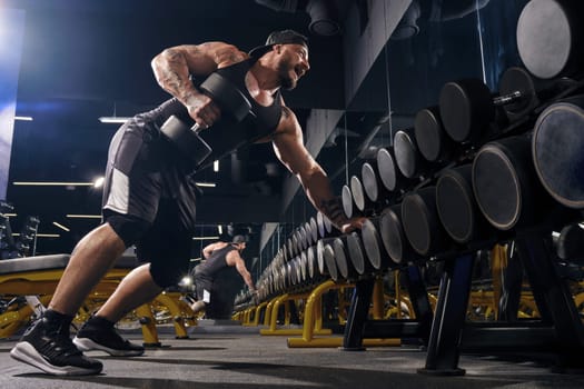 Attractive, tattooed, bearded muscular male in black shorts, vest, cap. Exercising with dumbbells for training triceps, leaning on set of black weights. Dark gym. Sport, fitness. Close up, side view