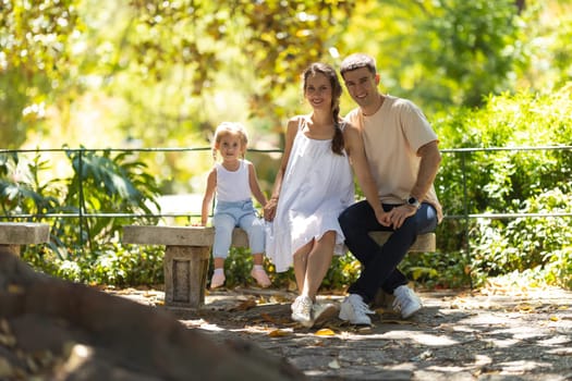 Family in park - a little girl with her parents sitting on a bench and looking in the camera. Mid shot