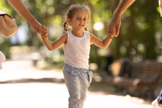 A little smiling girl walking in the park holding her parents by hands. Mid shot