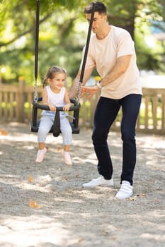 A man riding his daughter on the swings in the summer park. Mid shot