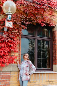 a beautiful girl stands against the background of the window of an old European house, entwined with a floating red color in autumn. Tourism & Travel Concept. Nice portrait of a young woman, in boho style outdoors in fall autumn day. Caucasian female girl 20 30 years old