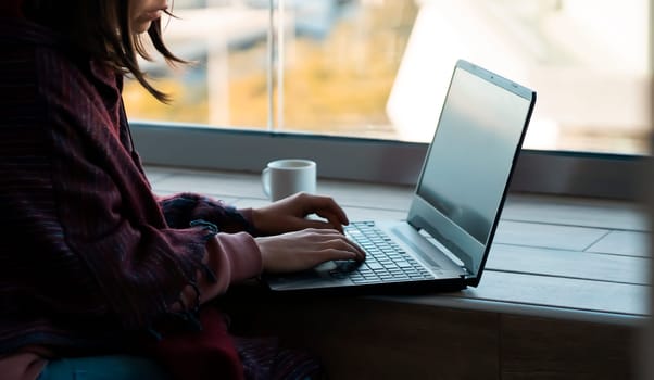 A young girl in a poncho sits on the terrace of her cozy house and works at a laptop with a cup of coffee, female hands are typing on the laptop keyboard, a woman works, studies online at sunset.