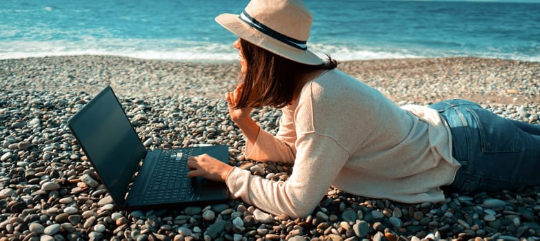 A young girl in a light hat and casual sweater lies on the beach by the sea with a laptop on a sunny day, works, studies, buys tickets during trip, a woman rests on vacation and types on the keyboard.