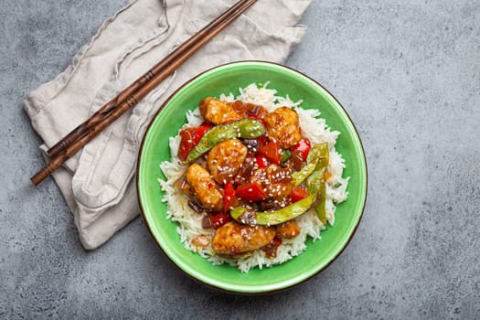 Asian sweet and sour sticky chicken with vegetables stir-fry and rice in ceramic bowl with chopsticks top view on gray rustic stone background, traditional Asian dish