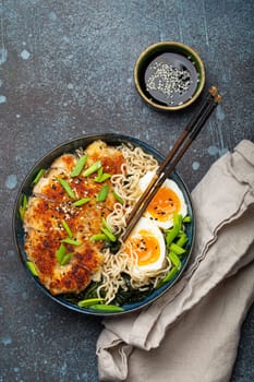 Asian noodles ramen soup with deep fried panko chicken fillet and boiled eggs in ceramic bowl with chop sticks and soy sauce on stone rustic background top view