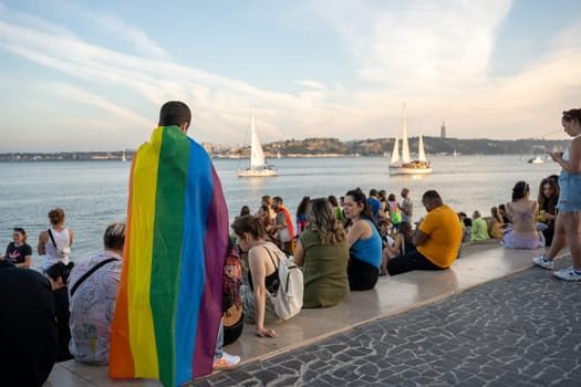 Lisbon, Portugal. 17 June 2023 Man with rainbow flag gay pride parade at promenade. Male gay standing outdoor during pride month. LGBT community wearing rainbow flag looking at yacht on water