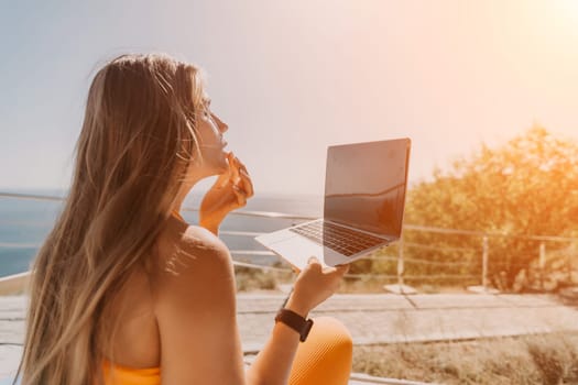 A woman with long blonde hair is sitting on a balcony and using a laptop. The sky is blue and the sun is shining. The woman is wearing a yellow shirt and a pair of sunglasses.