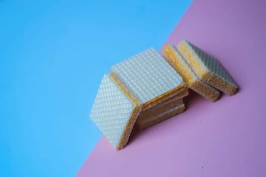 A pile of square, yellow wafers arranged on a blue and pink background. Copy space