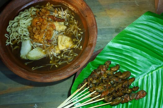 A bowl of Lontong Mie, a popular Indonesian soup made with rice cake, noodles, and various toppings, is served with a side of grilled satay, made from skewered chicken hearts, on a banana leaf in Surabaya.