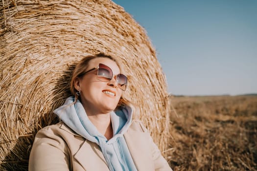 A woman wearing sunglasses and a beige jacket leans against a hay bale, smiling warmly.