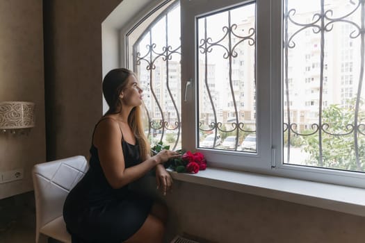 A woman in a black dress is sitting in front of a window with a bouquet of red roses