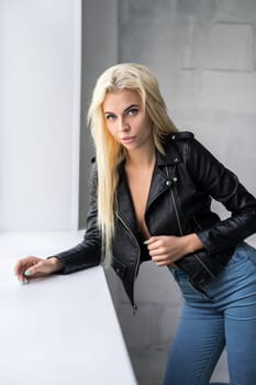 Young pretty slim blonde wearing black jacket and blue jeans staying near window sill in studio