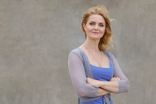 A beautiful woman in a good mood in a blue dress stands near the wall crossed, folded arms and looks at the camera.