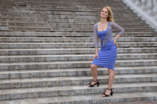 A pretty woman in a blue dress with beautiful hair, stands on the stairs. Portrait in full height.