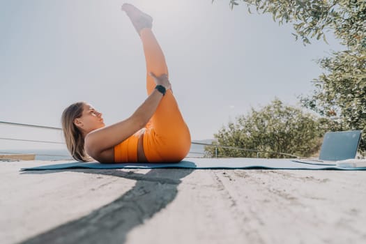A woman in orange activewear stretches her leg on a yoga mat outdoors with a laptop in the background and a clear sky.