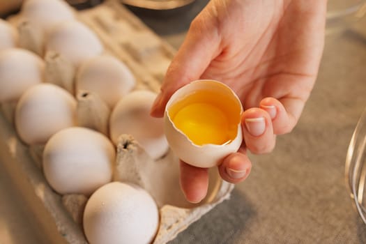 A person holding a cracked eggshell with a bright yellow yolk, surrounded by a tray of whole eggs. The setting is a kitchen with a rustic feel, featuring wooden elements and soft lighting.