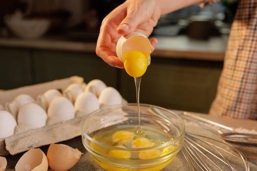 A person cracking an egg into a glass bowl, with several eggs in an egg carton in the background. The scene is set in a kitchen, showcasing the process of preparing food.