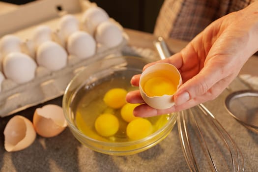 A woman's hand holding a broken chicken egg close-up over a bowl of raw eggs. There are several yolks in the bowl and more eggs in a cardboard box in the background.