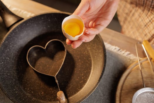 A person holding an egg with a broken shell, ready to crack it into a heart-shaped mold in a frying pan. The kitchen setting is warm and inviting, with utensils nearby.