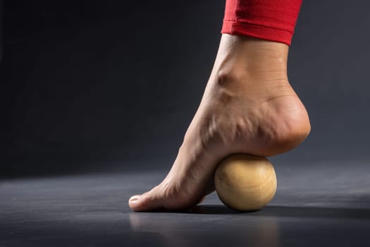 Myofascial release of foot muscles with a massage ball on a dark background, close-up. Concept on the topic of preventing foot fatigue after wearing high-heeled shoes.