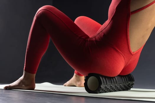 woman massaging gluteal muscles with rubber roller on floor, on dark background, healthy body concept and muscle tone