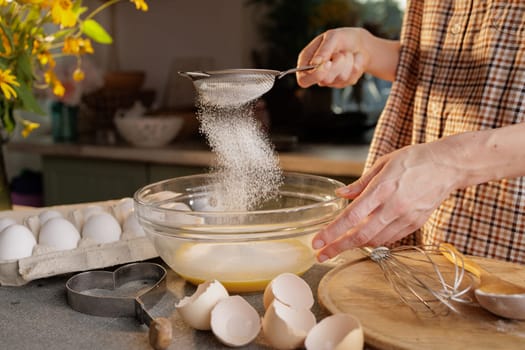 Close-up of female hands sifting flour and preparing dough in a transparent bowl against the background of a kitchen. Eggs in a box and a tool for making dough are on the table