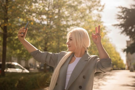 A woman is taking a picture of herself with her cell phone. She is wearing a gray jacket and scarf. The scene is set in a city with trees and cars in the background