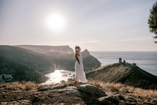 woman stands on a rocky hill overlooking a body of water. She is wearing a white dress and she is enjoying the view