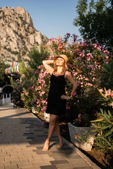 A woman in a black dress and straw hat stands on a path, posing with her hand on her hip, in front of a large, rocky cliff. family vacation concept