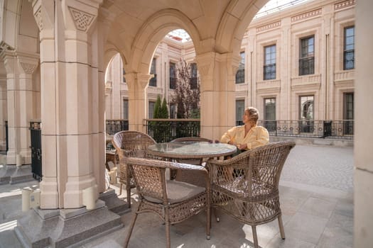A man sits alone at a table with wicker chairs under an arched stone structure.