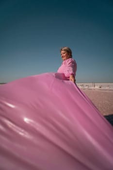 A woman in a pink dress stands on a beach, holding a large piece of pink fabric that billows in the wind. The sky is blue.