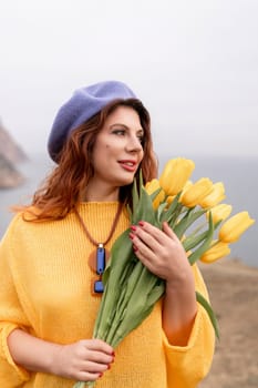A woman in a yellow sweater holding a bouquet of yellow tulips. She is wearing a blue hat and a necklace