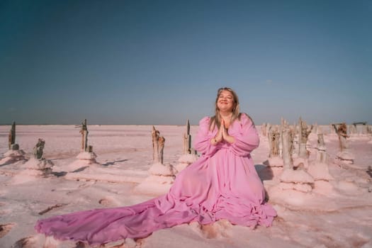 A woman in a pink dress sits on a salt flat, her hands clasped in prayer, surrounded by numerous salt formations. The sky is a pale blue.